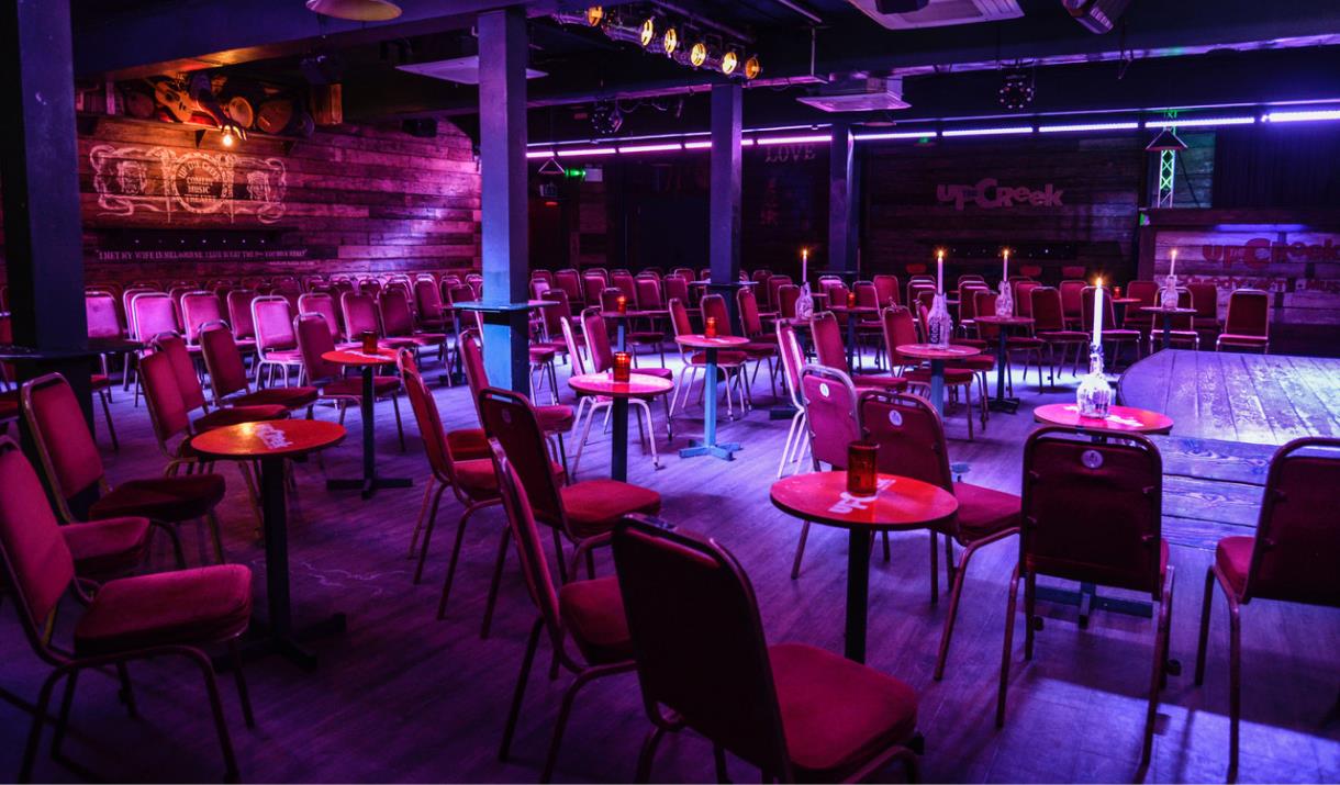 A picture taken inside Up the Creek Comedy Club. In the picture you can see lots of tables and chairs scattered around the room facing a small stage.