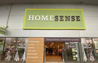 Outside Homesense in Charlton. A large silver building with a green, white and black sign at the top. There is also a row of windows showing all of th