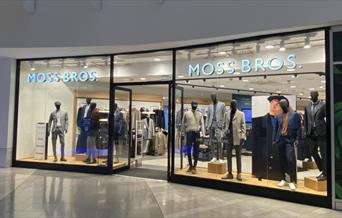 Outside Moss Bros at The O2. A modern shop with a brilliant window display, showing multiple mannequins wearing suits.