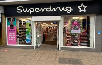 A photo taken outside of Superdrug, Woolwich. Showing a black and white modern shop with a wide range in products.