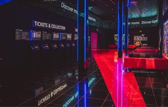 A well taken photo of inside of Cineworld O2, in the picure you can see the tickets and collection area, red and black sofas, movie posters and the en