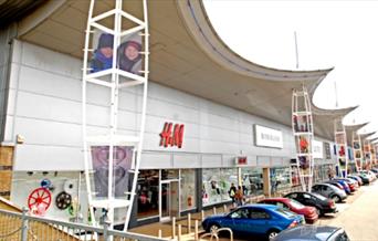 A picture of Greenwich Shopping park, showing a long row of shops. Right in front of them is the car park for easy access.