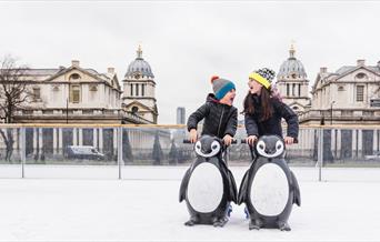 Two children laughing as they hold onto model penguins to steady themselves on the ice at the Queen's House Ice Rink in Greenwich.
