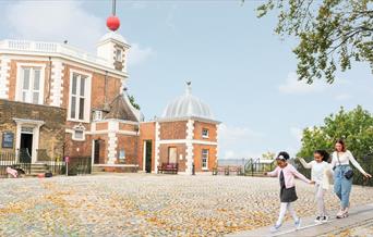 A photo outside of the Royal observatory entrance, there is blue skies, trees and a blanket of orange leaves spread across the stone pavement. Too the