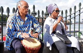 Photo of a man in a blue and white printed shirt sitting playing a drum next to a woman sitting beside him singing into a microphone wearing a lilac h