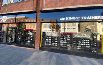 Outside JD Sports Eltham, showing a metallic black shop front presenting a wide range of products.