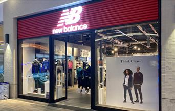 Outside New Balance at The O2. A stylish shopfront with a red banner and windows showing off all their great products.