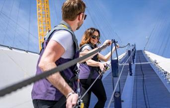 A couple climb the blue walkway of Up at The O2 on Greenwich Peninsula on a sunny, blue-skied day.