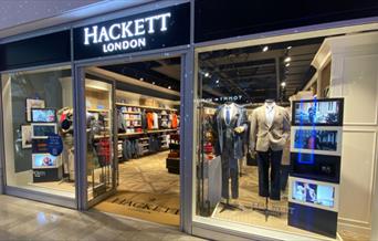 Outside Hackett inside The O2. A modern and welcoming shop front with open doors and windows presenting a range of clothing.
