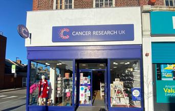 Outside Cancer Research UK, Eltham. A blue and white shop with a cancer research banner at the top. In the windows is a huge selection of items availa