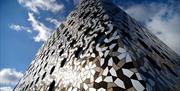 Exterior image of Ravensbourne University London with the building swathed in different coloured, tesellated aluminium tiles and circular windows of d