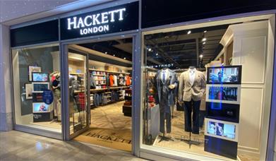 Outside Hackett inside The O2. A modern and welcoming shop front with open doors and windows presenting a range of clothing.