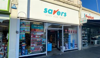 Outside Savers in Eltham. A white shop with a blue and red logo and a selection of products in the window.