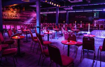 A picture taken inside Up the Creek Comedy Club. In the picture you can see lots of tables and chairs scattered around the room facing a small stage.