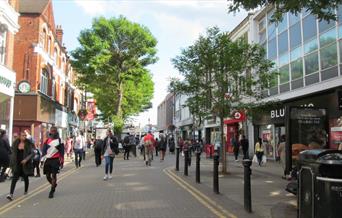 Woolwich high street, a road with shops lined up on each side. Also showing benches and bins for public use.