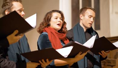 Two men and woman - wearing a red scarf - hold hymn books and sing as if in a choir.