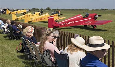 Row of people watching small yellow and red aircraft about to take off