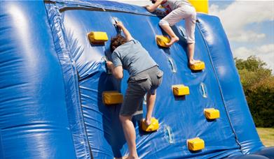 Two people climbing up a blue inflatable wall in a park