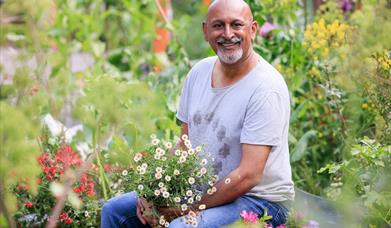 Manoj Malde Sitting on a bench, surrounded by green foliage and some red plants. He's holding some white daisies