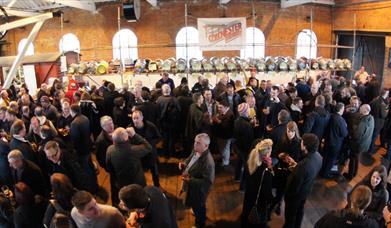 A crowd of people, stood in front of kegs of beer.