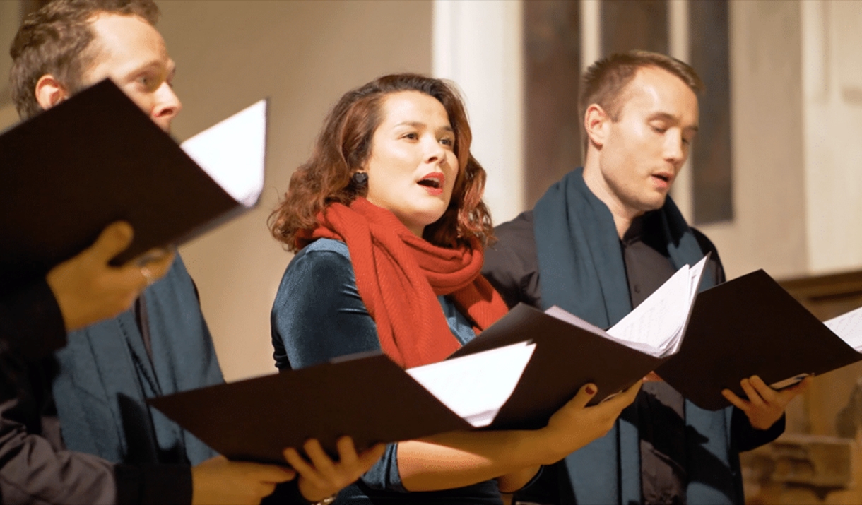 Two men and woman - wearing a red scarf - hold hymn books and sing as if in a choir.