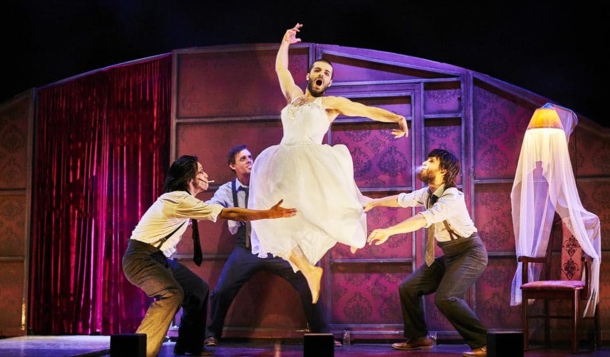 A man in a wedding dress jumps in the style of a ballet dancer. Three other besuited men act as if to catch him.