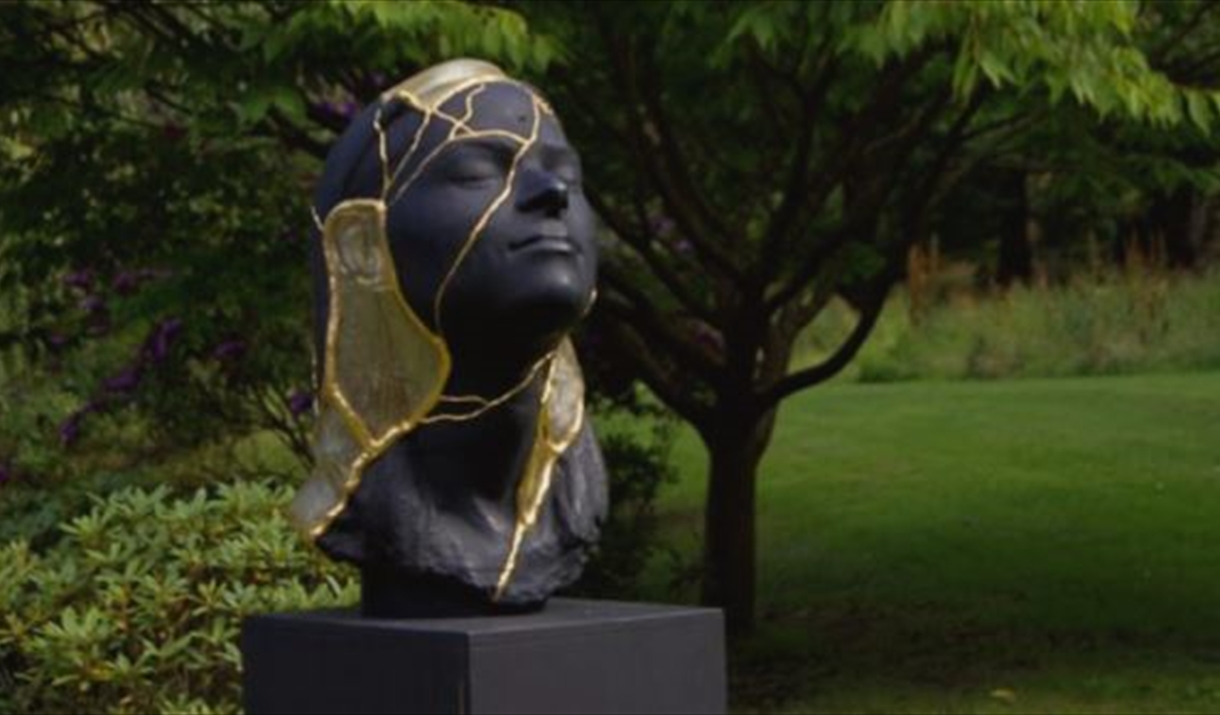 Black sculpture of a human face with gold leaf accents, set on a lawn.