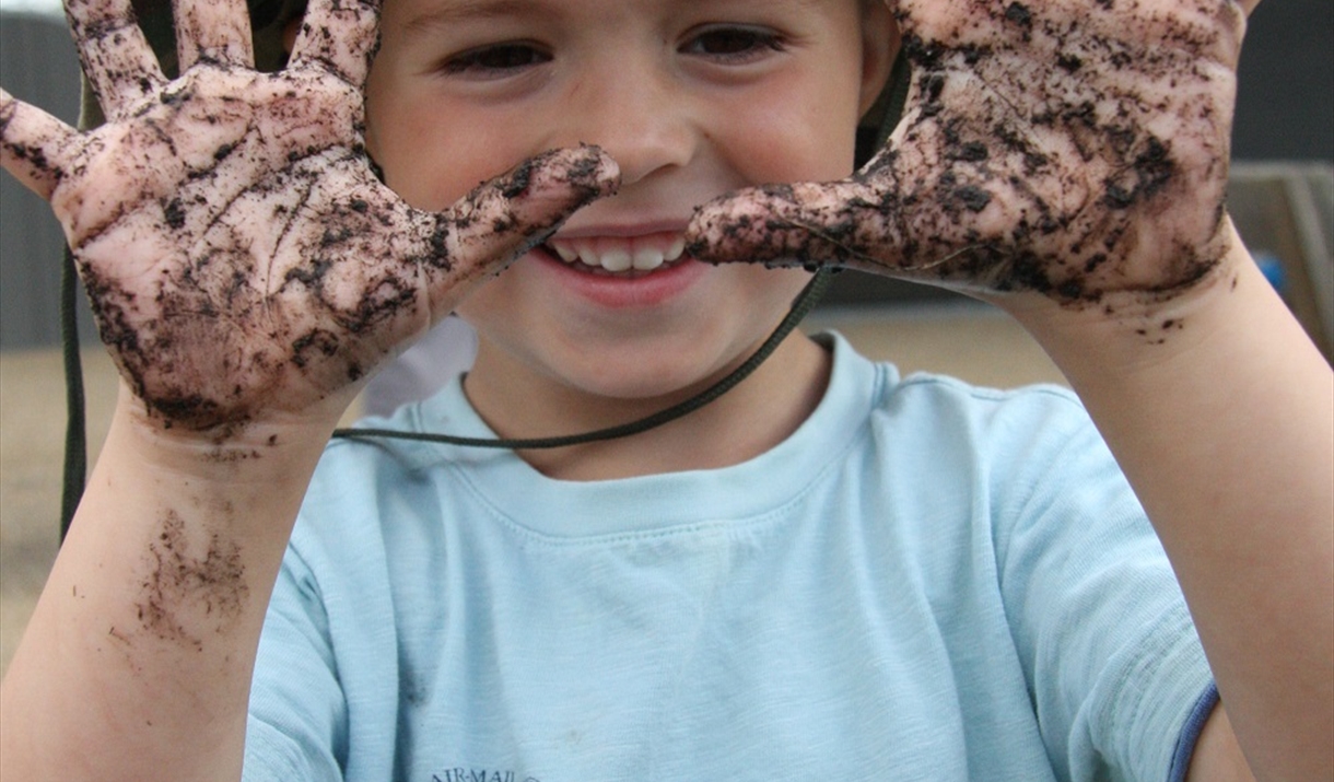 A young boy in a blue shirt and green sun hat holds up his very muddy hands, with a big grin on his face. Picture by Louise Moss