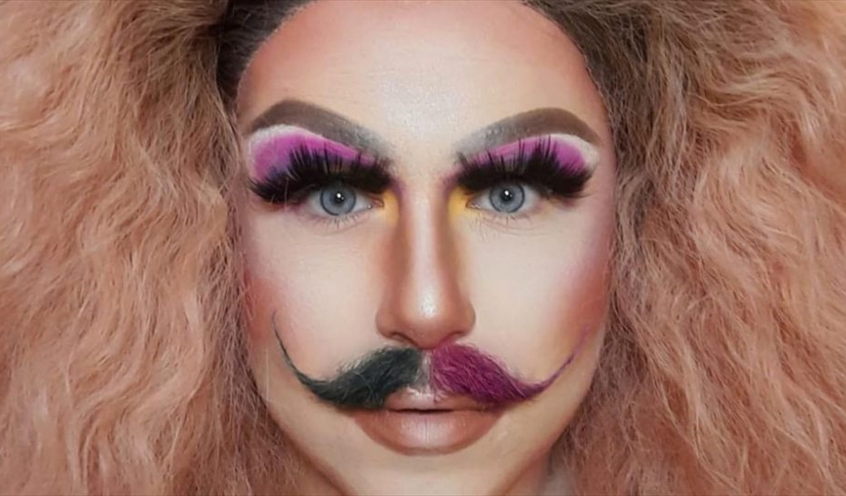 A drag artist with wild blonde hair wearing false eyelashes, pink eyeshadow and a mustache