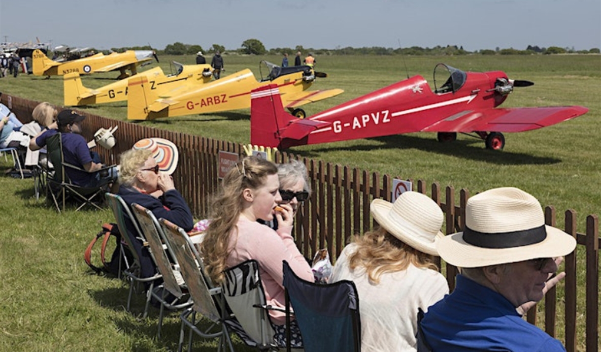 Row of people watching small yellow and red aircraft about to take off
