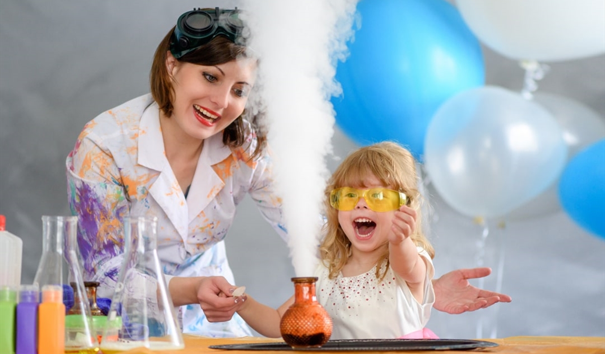 A woman, dressed in a lab coat splattered with paint demonstrates steam coming from a beaker to an excited looking child.