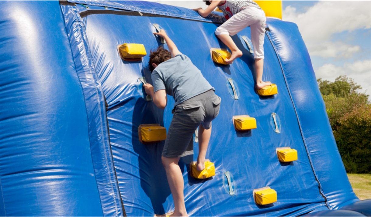 Two people climbing up a blue inflatable wall in a park