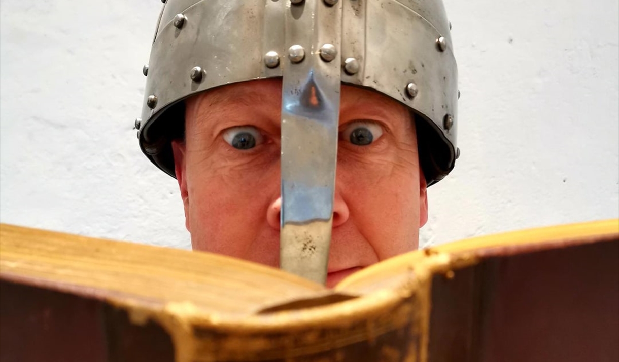 A man wearing a knight's helmet reads from a book