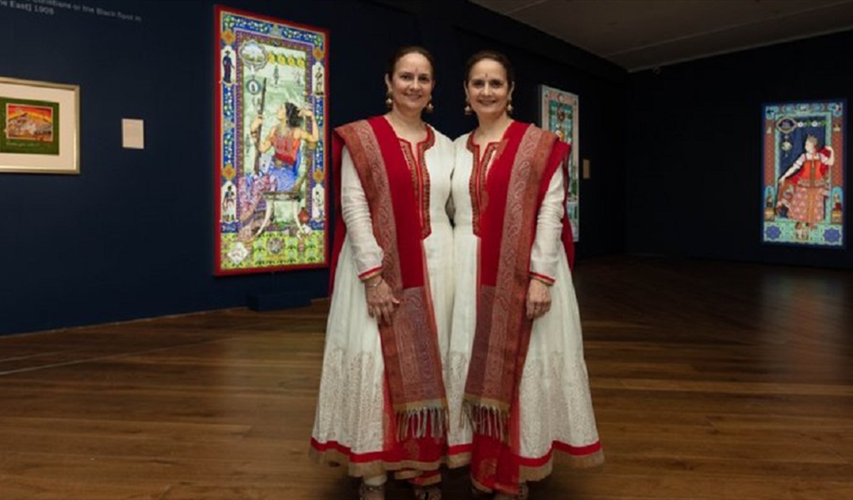 Two women in traditional Indian costume stand next to each other giving a somewhat symmetrical appearance. Illuminated artworks can be seen in the bac