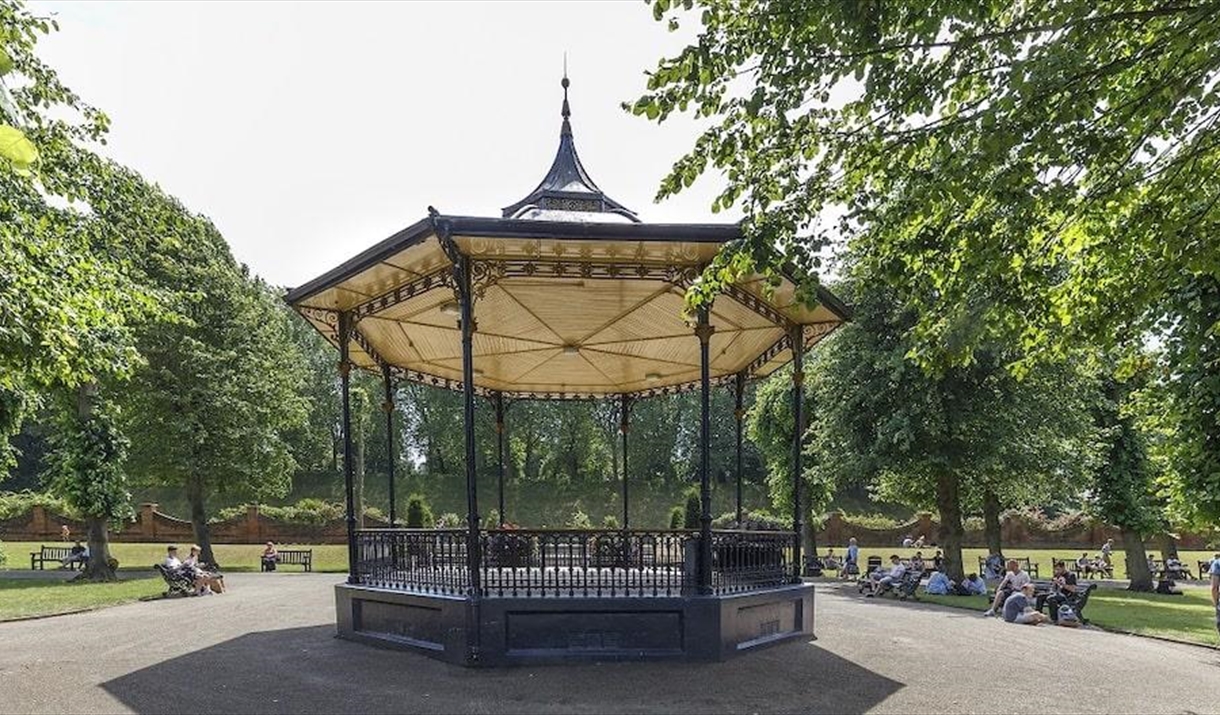 the victorian bandstand in castle park, surrounded by trees and people sitting on wooden benches. the bandstand has dark blue decorative ironwork and