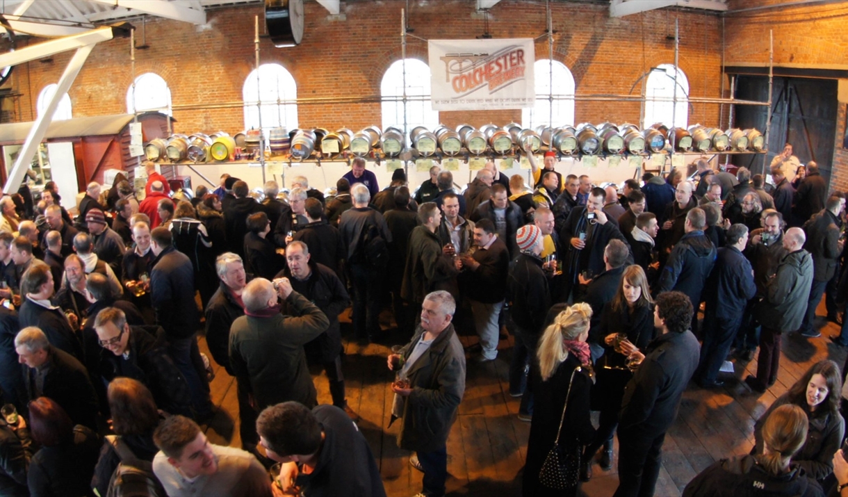 A crowd of people, stood in front of kegs of beer.