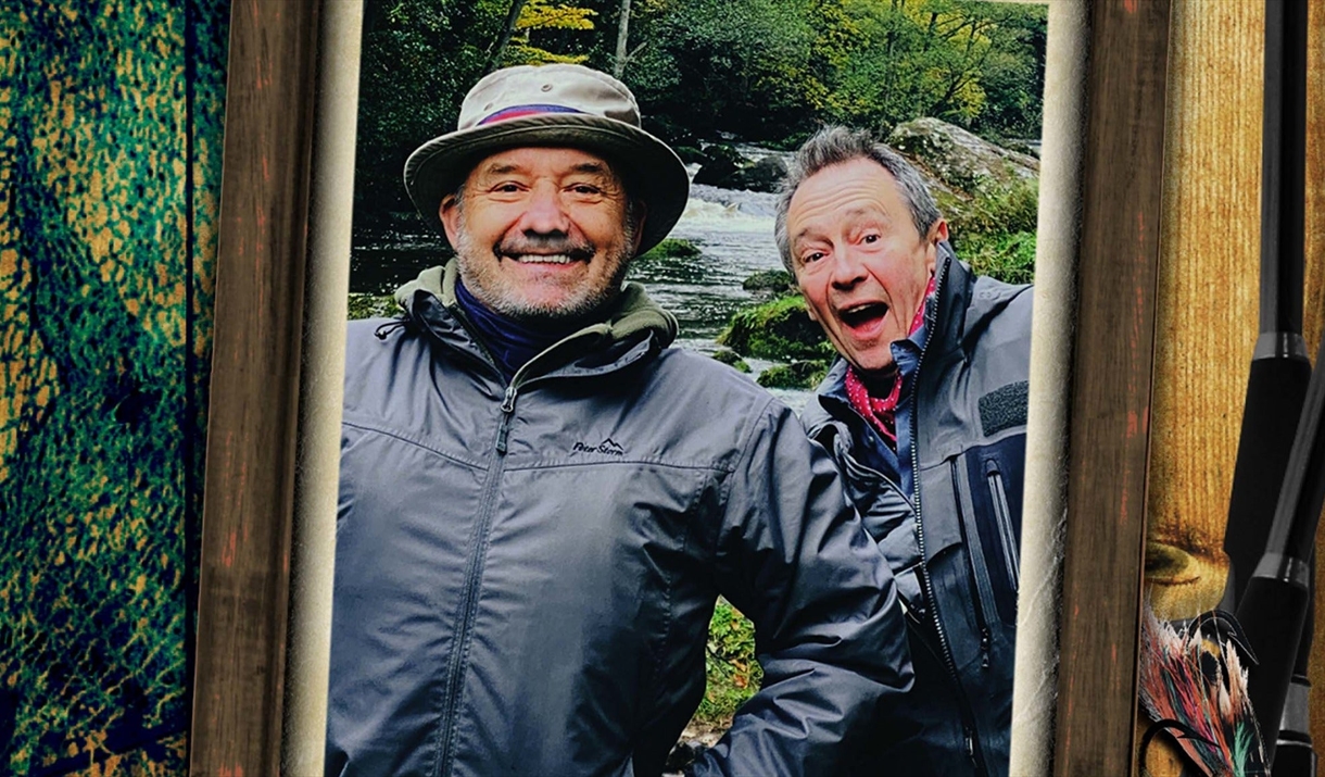 Bob and Paul stand in wet weather gear in front of a river