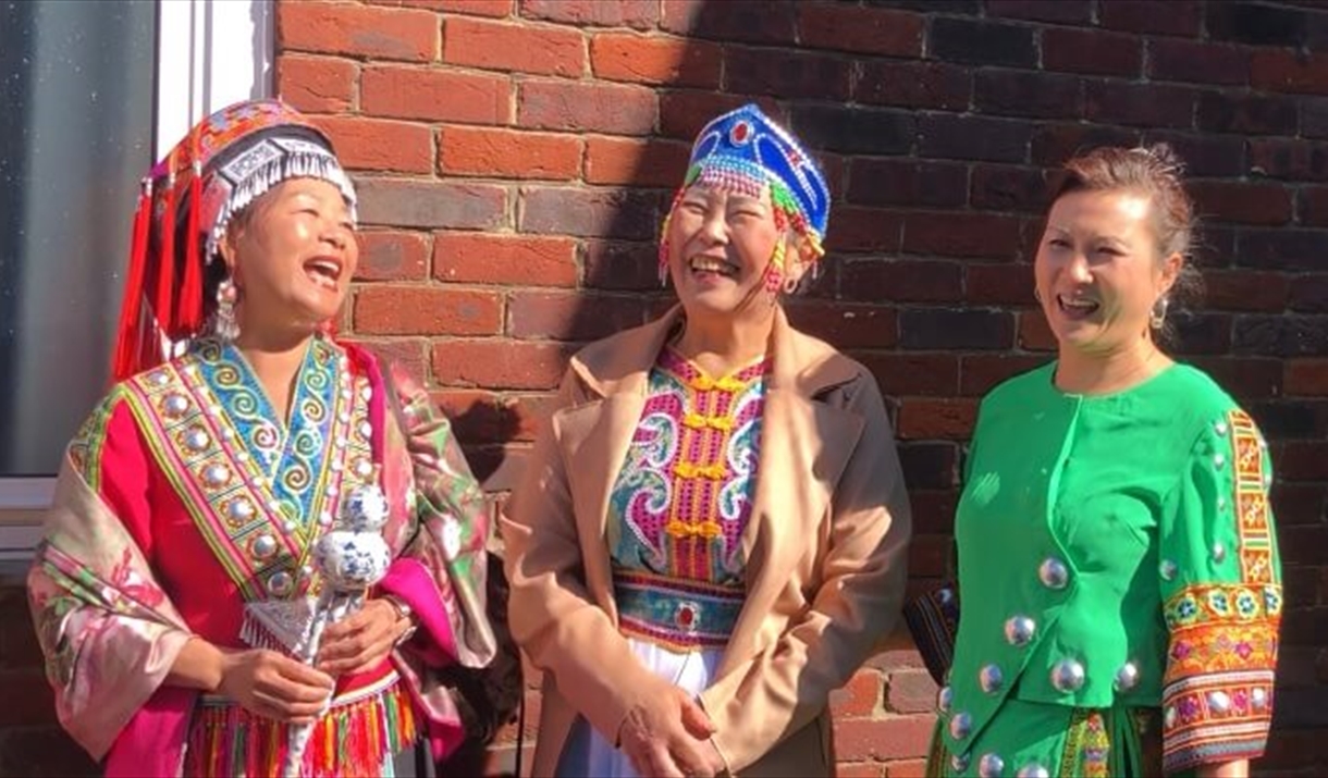 three women standing with their backs against a brick wall, the sun is shining and they are laughing, they are dressed in brightly coloured traditiona