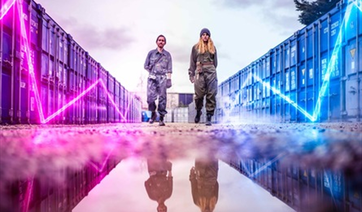 Two youths in boiler suits stand between shipping containers. Neon lighting zig-zags on the side of the containers.