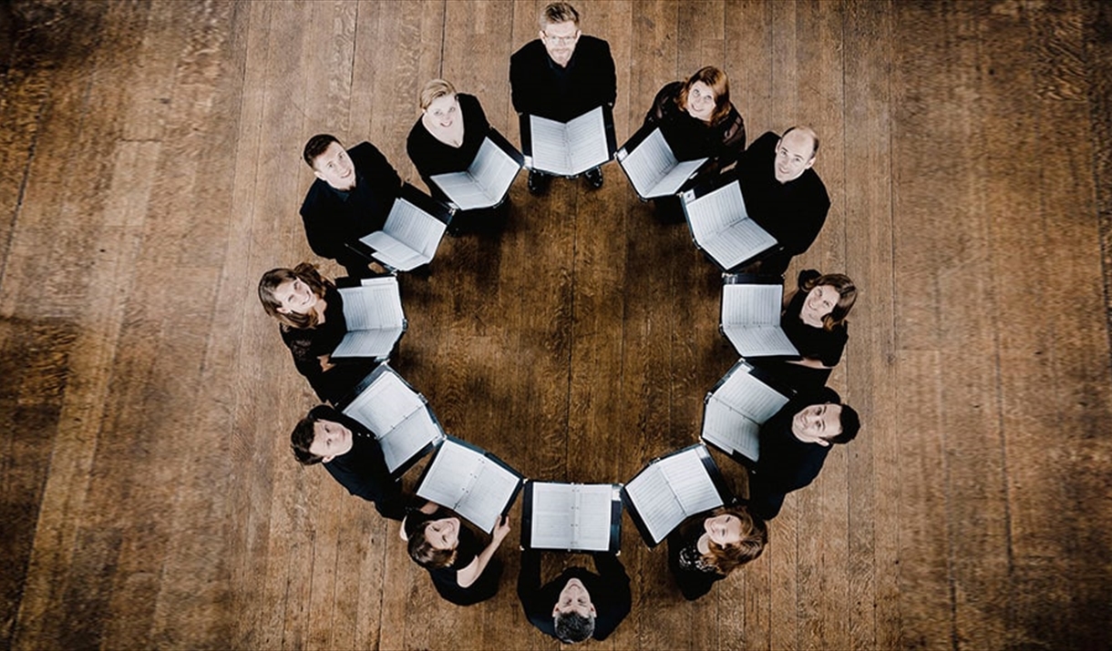 A choir, photographed from above, stand in a circle with their sheet music open.