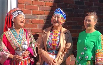 three women standing with their backs against a brick wall, the sun is shining and they are laughing, they are dressed in brightly coloured traditiona