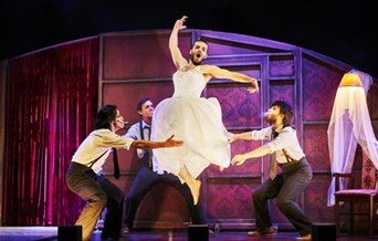 A man in a wedding dress jumps in the style of a ballet dancer. Three other besuited men act as if to catch him.