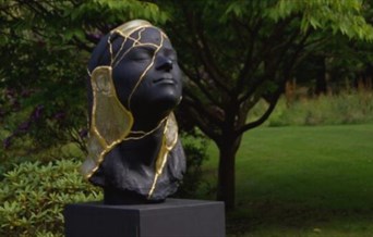 Black sculpture of a human face with gold leaf accents, set on a lawn.