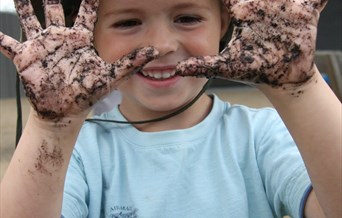 A young boy in a blue shirt and green sun hat holds up his very muddy hands, with a big grin on his face. Picture by Louise Moss