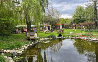 Pond surrounded by picnic benches, willow trees, and the pink Galloping Grind horsebox café in the background