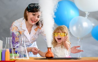 A woman, dressed in a lab coat splattered with paint demonstrates steam coming from a beaker to an excited looking child.