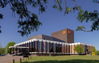 Photo of the Queen's Theatre Hornchurch building against a blue sky. The building has glass windows from floor to ceiling on all 3 front sides.