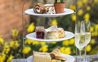 Close-up of a three-tier cake stand showing an afternoon tea; sandwiches/savoury pastries, scones with cream and jam, and a selection of cake.