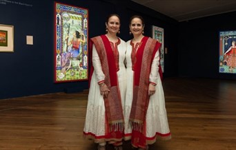 Two women in traditional Indian costume stand next to each other giving a somewhat symmetrical appearance. Illuminated artworks can be seen in the bac