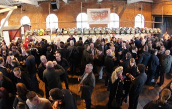 A crowd of people, stood in front of kegs of beer.